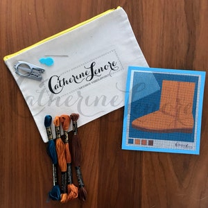 May include: A needlepoint kit with a blue canvas, a brown boot design, and four skeins of embroidery floss in blue, orange, and brown. The kit comes in a white canvas bag with the text "Catherine Lenore Modern Needlepoint" printed on it.