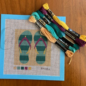 May include: A blue canvas with a white grid pattern and a design of two green and purple flip flops. The canvas is surrounded by a light blue border. There are six skeins of embroidery floss in various colors: yellow, orange, green, teal, and purple.