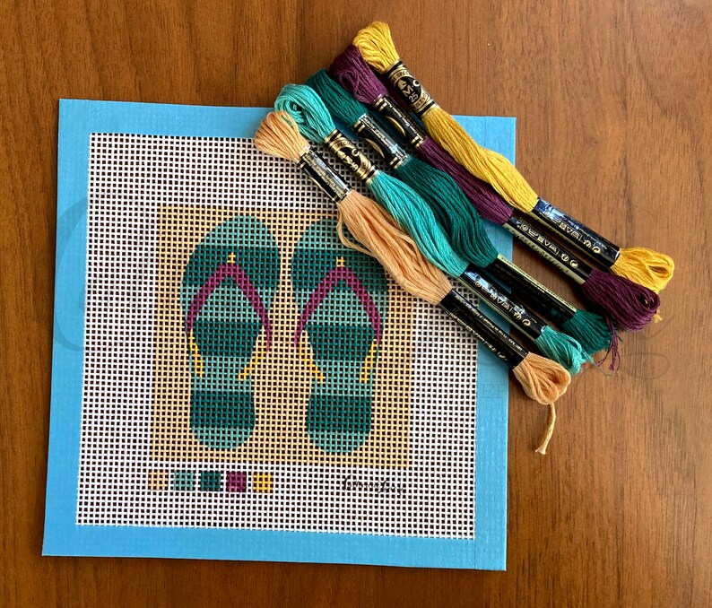 May include: A blue canvas with a white grid pattern and a design of two green and purple flip flops. The canvas is surrounded by a light blue border. There are six skeins of embroidery floss in various colors: yellow, orange, green, teal, and purple.