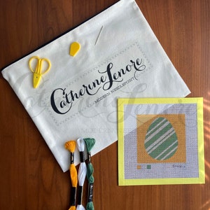 May include: A needlepoint kit with a yellow canvas, green and yellow embroidery floss, and a needlepoint pattern of a green and white striped egg on a yellow background. The kit is packaged in a white canvas bag with the text "Catherine Lenore Modern Needlepoint" printed on it.