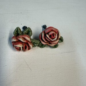 May include: Two ceramic drawer knobs shaped like blooming roses. Each knob features layered petals in shades of pink and white with dark green leaves at the base. A small black metal screw protrudes from the back of each floral piece.