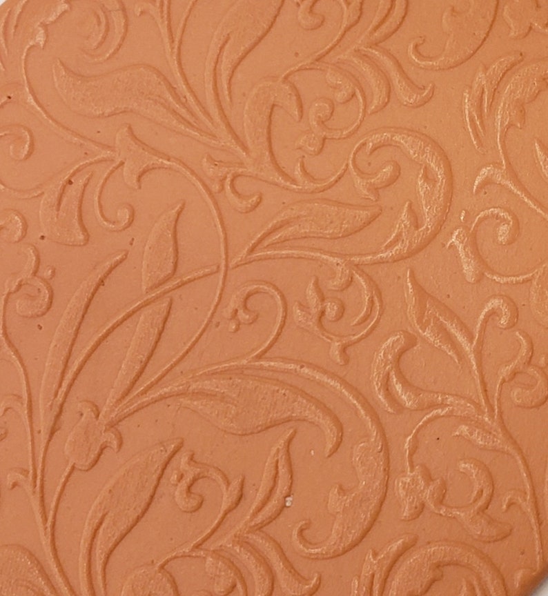 May include: A close-up of a terracotta-colored tile with an embossed floral pattern. The pattern is intricate and detailed, with swirling leaves and vines.
