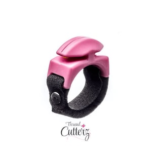 May include: A pink thread cutter ring with a black adjustable strap. The cutter is designed to be worn on the finger and has a sharp blade for cutting thread.