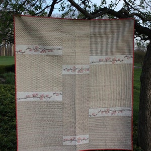 May include: A beige and white patchwork quilt with a red border and embroidered pink flowers. The quilt is hanging from a tree branch.
