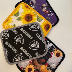 May include: Four different pot holders with various designs. One is purple with a black and white design, one is yellow with sunflowers, one is black with the word "RAIDERS" repeated, and one is black and white with chickens and sunflowers.