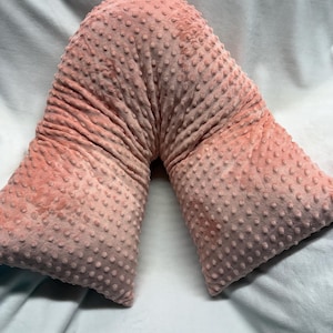 May include: A V-shaped, pink pillow with a textured, dotted surface. The pillow is made of a soft material and is designed for comfort and support. The pillow's color is a muted, dusty rose.