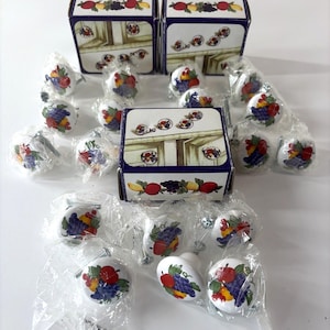 May include: A collection of round white ceramic cabinet knobs featuring a colourful fruit pattern of grapes, apples, and pears. The knobs are individually wrapped in clear plastic and are displayed alongside three matching cardboard boxes.