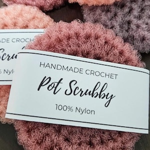 Könnte beinhalten: Handgefertigte Häkel-Topf-Schrubbe in staubigem Rosaton. Die Schrubbe besteht aus 100 % Nylon und hat ein Etikett mit der Aufschrift "Handmade Crochet Pot Scrubby 100% Nylon".
