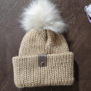 May include: A tan knit beanie with a large, fluffy, cream-colored pom-pom on top. The hat features a folded brim and a small, brown leather tag with the text "RURAL CURIO" attached to the front.