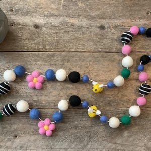 May include: A colorful felt garland with white, black, blue, green, pink, and yellow felt balls. The garland features felt flowers and bees.
