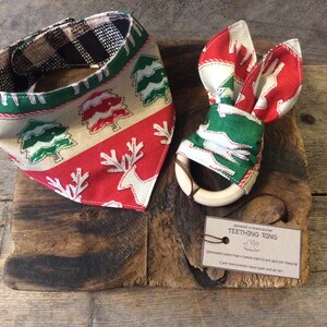 May include: A Christmas-themed baby bandana and teething ring set. The bandana is red, green, and white with a pattern of reindeer and Christmas trees. The teething ring is made of wood and is wrapped in the same fabric as the bandana.