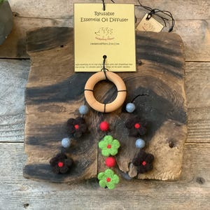 May include: A reusable essential oil diffuser with a wooden ring and felt flower and ball accents. The diffuser hangs from a wooden plaque. The flowers are brown and green with red centers, and the balls are gray. The tag reads "Reusable Essential Oil Diffuser".