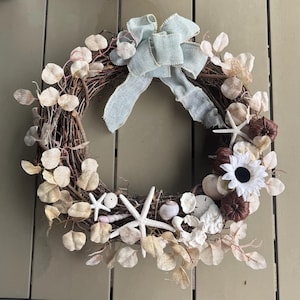 May include: A circular wreath made from twigs, decorated with dried leaves, seashells, starfish, and small brown pumpkins. A light blue burlap bow is at the top. The wreath is a decorative item.