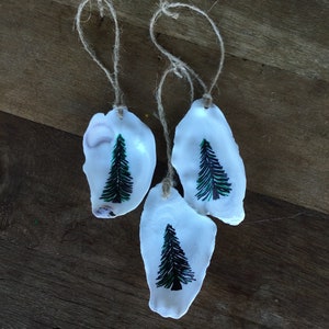 May include: Three white oyster shells with green and black painted pine trees, each with a twine loop for hanging.
