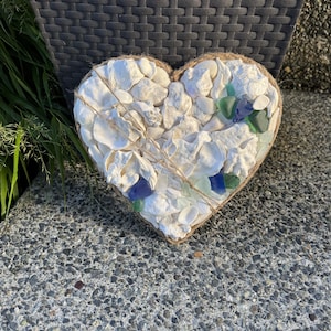 May include: A heart-shaped wall hanging crafted from white seashells and scattered pieces of blue and green sea glass. The arrangement is bordered by a natural jute twine rope, creating a textured surface with a variety of shell shapes.