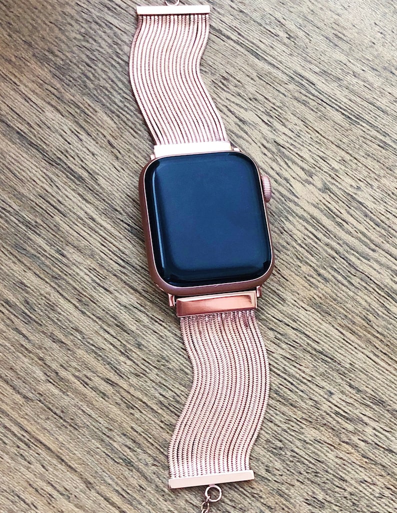 May include: A rose gold Apple Watch with a rose gold metal band. The band is made of a series of thin, linked metal strips.