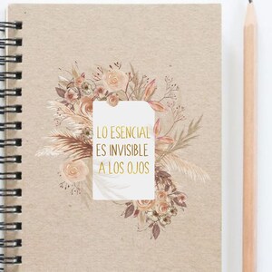 Op de afbeelding: Een spiraalgebonden notitieboek met een beige kaft. De kaft heeft een bloemenontwerp in tinten perzik, bruin en beige, met de tekst "LO ESENCIAL ES INVISIBLE A LOS OJOS" in gouden letters. Een houten potlood ligt naast het notitieboek.