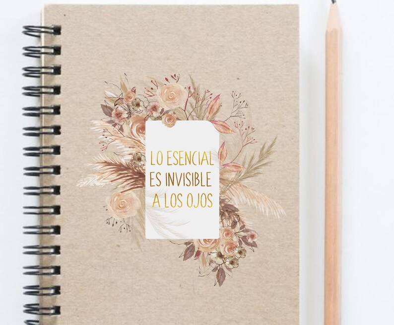 Op de afbeelding: Een spiraalgebonden notitieboek met een beige kaft. De kaft heeft een bloemenontwerp in tinten perzik, bruin en beige, met de tekst "LO ESENCIAL ES INVISIBLE A LOS OJOS" in gouden letters. Een houten potlood ligt naast het notitieboek.