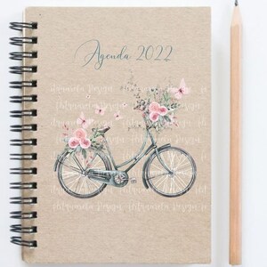 Pu&ograve; includere: Un'agenda con una copertina in carta marrone e una rilegatura a spirale. La copertina presenta un'illustrazione ad acquerello di una bicicletta con un cestino di fiori rosa e farfalle. Il testo "Agenda 2022" &egrave; scritto in corsivo nella parte superiore della copertina.
