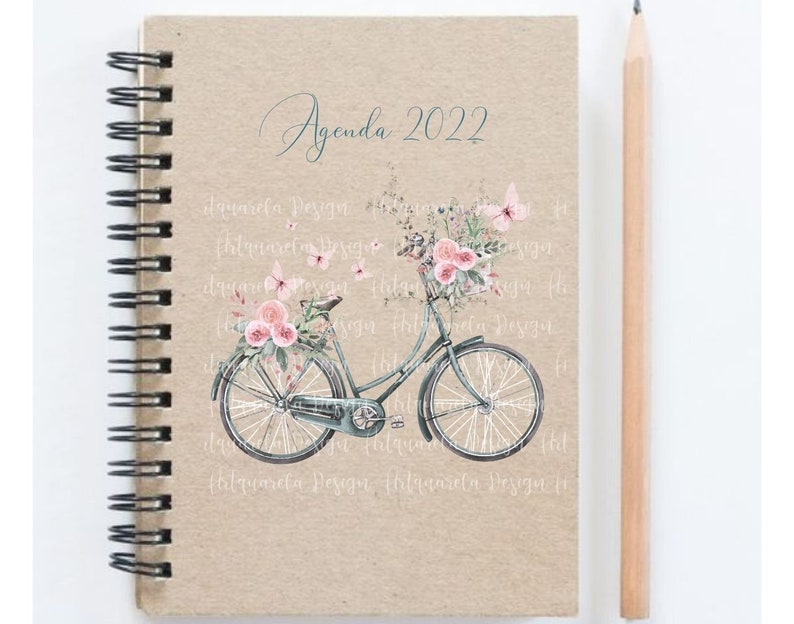 Pu&ograve; includere: Un'agenda con una copertina in carta marrone e una rilegatura a spirale. La copertina presenta un'illustrazione ad acquerello di una bicicletta con un cestino di fiori rosa e farfalle. Il testo "Agenda 2022" &egrave; scritto in corsivo nella parte superiore della copertina.
