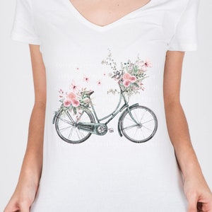 Pu&ograve; includere: Una maglietta bianca con una bicicletta verde con un cestino pieno di fiori rosa e foglie verdi. Ci sono farfalle che volano intorno alla bicicletta.
