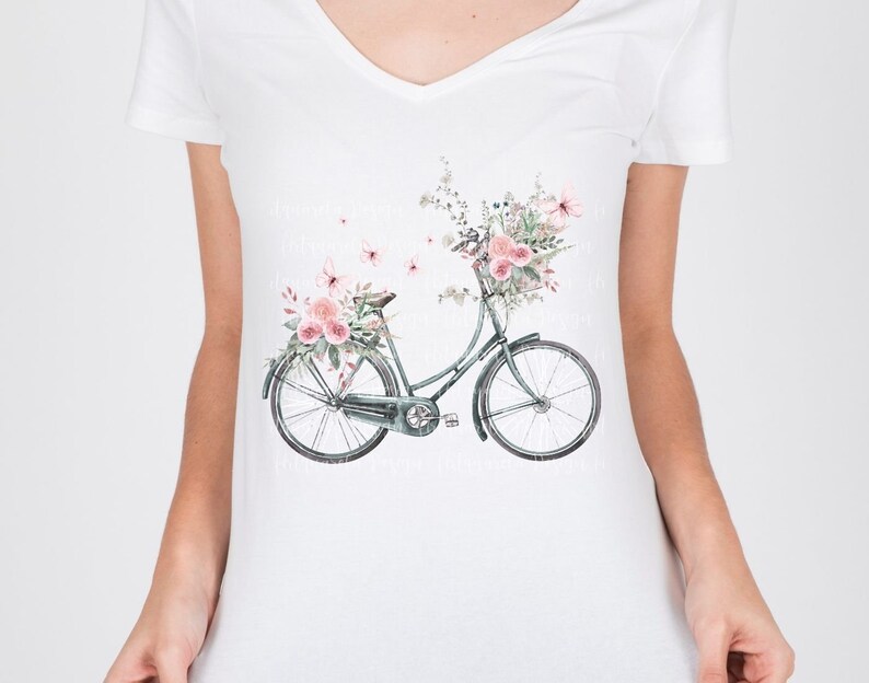 Pu&ograve; includere: Una maglietta bianca con una bicicletta verde con un cestino pieno di fiori rosa e foglie verdi. Ci sono farfalle che volano intorno alla bicicletta.