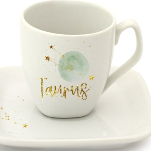 May include: White ceramic coffee mug with a gold glitter design of the Taurus constellation and the word "Taurus". The mug is paired with a matching saucer.