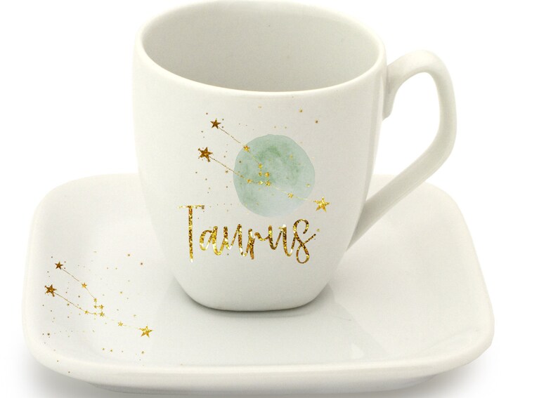 May include: White ceramic coffee mug with a gold glitter design of the Taurus constellation and the word "Taurus". The mug is paired with a matching saucer.