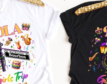 May include: Two t-shirts with a colourful graphic design featuring a street lamp, musical instruments, and the text "NOLA Girls Trip 2025". One shirt is white and the other is black.
