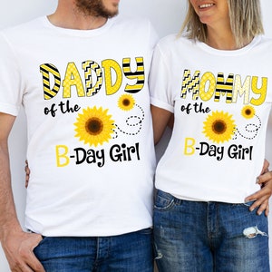 Peut inclure: Deux t-shirts blancs avec des motifs de tournesols assortis. L'un porte l'inscription "DADDY of the B-Day Girl" en lettres jaunes et noires, avec un motif de tournesol. L'autre dit "MOMMY of the B-Day Girl".