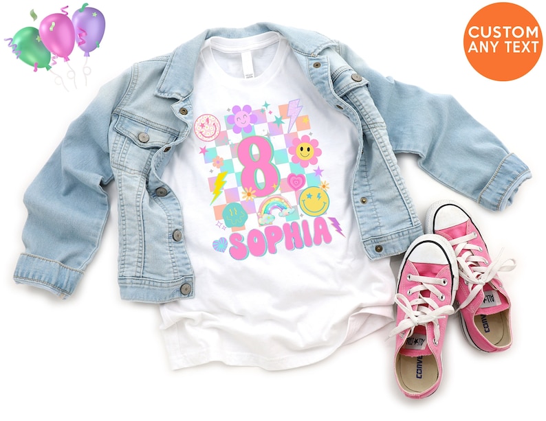 May include: A white t-shirt with a colourful graphic design featuring the number 8, a rainbow, smiley faces, and the name "Sophia" in pink. The shirt is paired with a light blue denim jacket and pink trainers. Balloons are in the upper left corner.