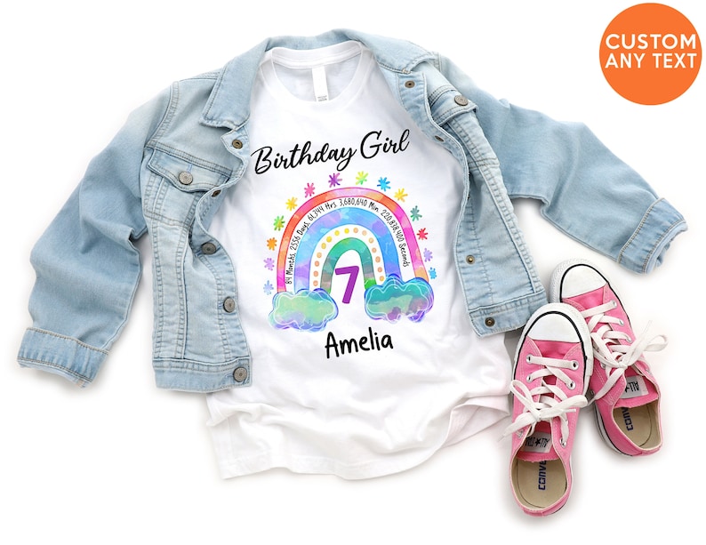 May include: White t-shirt with a rainbow design and the text "Birthday Girl". The rainbow has a number 7 in the center and the text "Amelia" below. The shirt is laid out on a white surface with a denim jacket and pink sneakers.