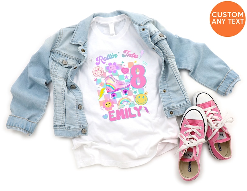 以下が含まれることがあります： 白いTシャツに、ピンクとブルーのローラースケート、虹、そして「Rollin' Into 8 Emily」というテキストのグラフィックが描かれています。