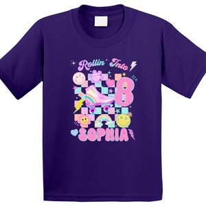 以下が含まれることがあります： 紫色のTシャツに、ピンクとブルーのローラースケートのグラフィック、虹、そして「Rollin' Into 8 Sophia」の文字がプリントされています。