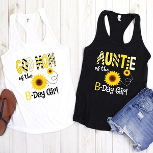 Peut inclure: Deux d&eacute;bardeurs, un blanc et un noir, avec des motifs de tournesols et du texte. Le d&eacute;bardeur blanc dit "GOD MOM of the B-Day Girl". Le d&eacute;bardeur noir dit "AUNTIE of the B-Day Girl". Des sandales et un short en jean sont &eacute;galement visibles.