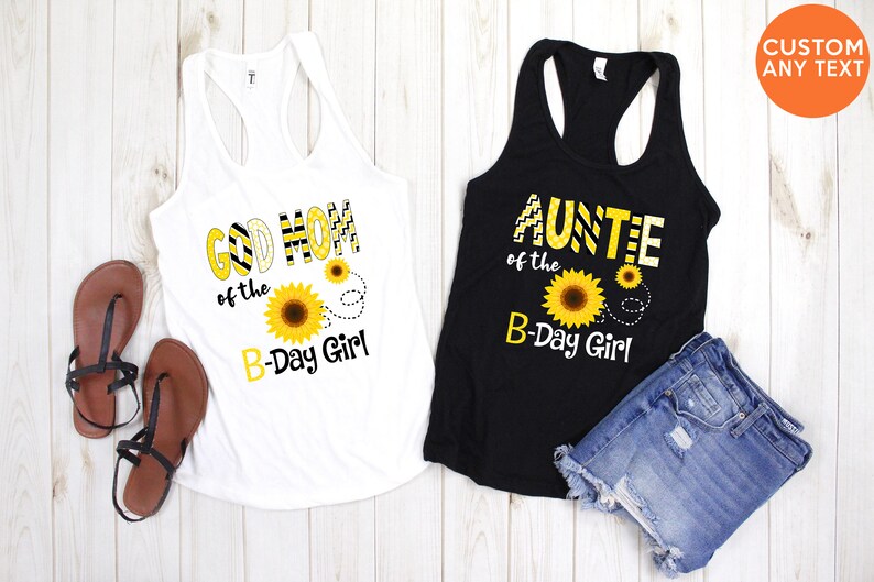 Peut inclure: Deux d&eacute;bardeurs, un blanc et un noir, avec des motifs de tournesols et du texte. Le d&eacute;bardeur blanc dit "GOD MOM of the B-Day Girl". Le d&eacute;bardeur noir dit "AUNTIE of the B-Day Girl". Des sandales et un short en jean sont &eacute;galement visibles.