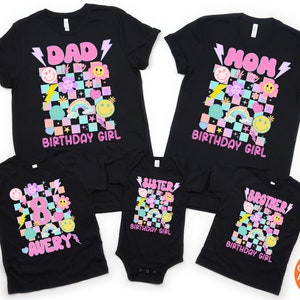 May include: Black t-shirts with colourful retro graphics. Designs include "DAD," "MOM," "8," "SISTER," and "BROTHER" with "BIRTHDAY GIRL" text. The shirts feature smiley faces, rainbows, and lightning bolts. The name "AVERY" is also displayed.
