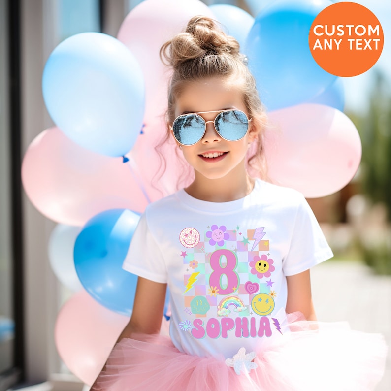 May include: A girl wearing a white t-shirt with a colourful graphic design featuring the number 8, smiley faces, and the name "Sophia". She is wearing sunglasses and a pink tutu, with blue and pink balloons in the background.