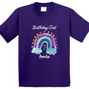 May include: Purple short-sleeved t-shirt with a rainbow design and the text "Birthday Girl" and "Amelia". The rainbow has a number 7 in the center and the text "81 Months 2358 Days 61,344 Hrs 3,680,540 Min 220,838,000 Seconds" around it.