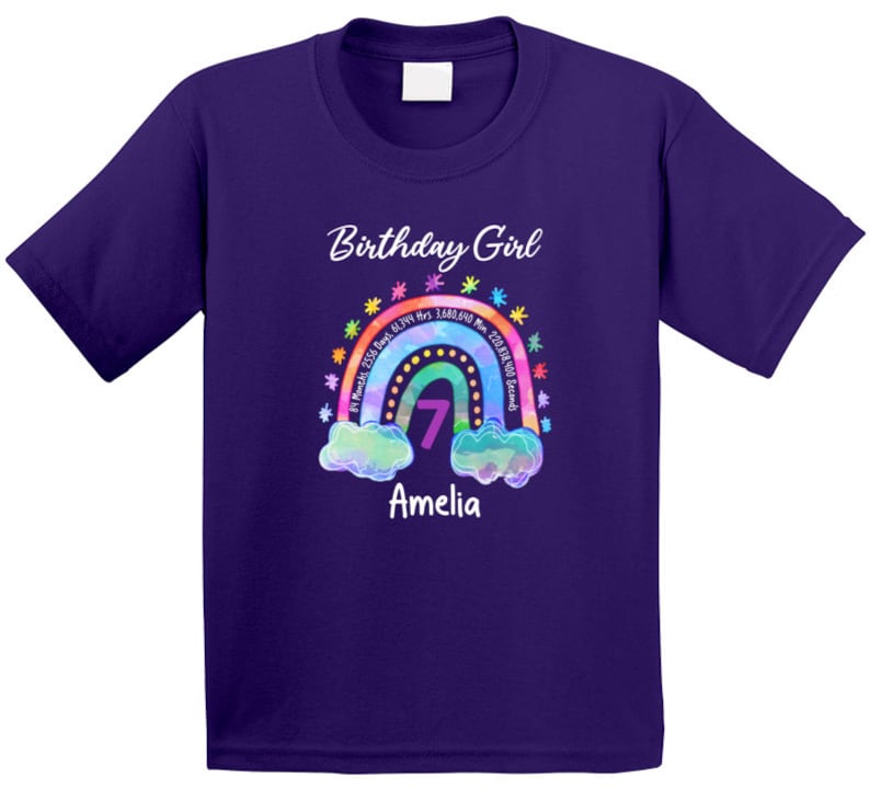 May include: Purple short-sleeved t-shirt with a rainbow design and the text "Birthday Girl" and "Amelia". The rainbow has a number 7 in the center and the text "81 Months 2358 Days 61,344 Hrs 3,680,540 Min 220,838,000 Seconds" around it.