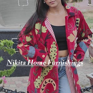 May include: A long, vibrant red velvet coat featuring intricate, multi-colored floral embroidery, a collared neckline, and a red and white striped inner lining. The text "Nikita Home Furnishings" is overlaid across the center of the garment.