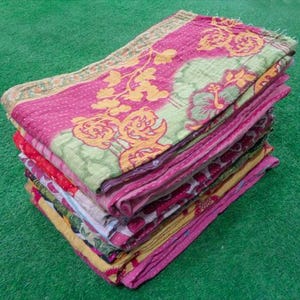 Pode incluir: Uma pilha de colchas dobradas e coloridas. A colcha superior apresenta um fundo rosa com padr&otilde;es florais amarelos e detalhes verdes. Outras colchas na pilha t&ecirc;m desenhos florais vermelhos, rosa e amarelos. As colchas est&atilde;o empilhadas sobre uma superf&iacute;cie verde.
