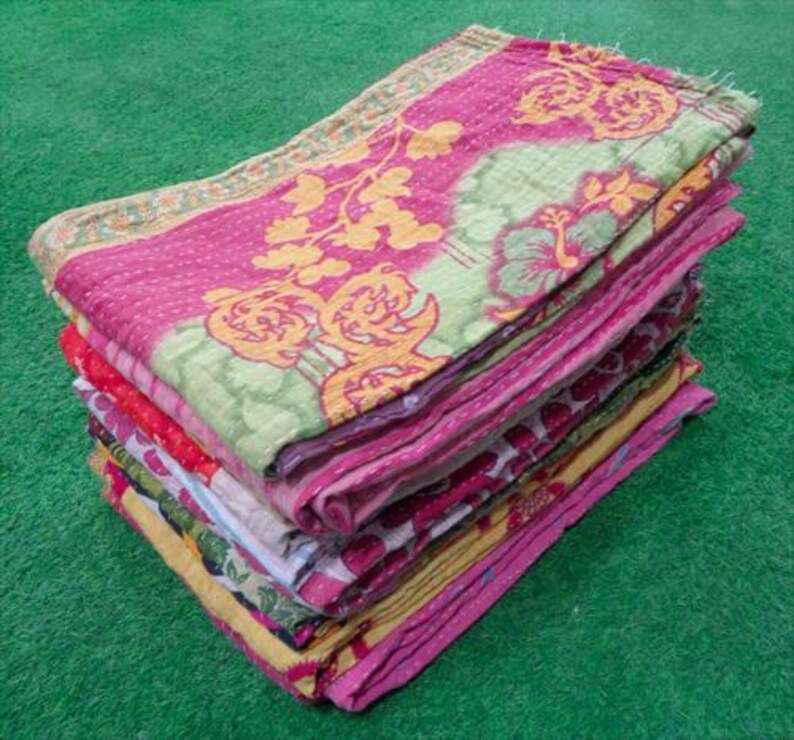 Pode incluir: Uma pilha de colchas dobradas e coloridas. A colcha superior apresenta um fundo rosa com padr&otilde;es florais amarelos e detalhes verdes. Outras colchas na pilha t&ecirc;m desenhos florais vermelhos, rosa e amarelos. As colchas est&atilde;o empilhadas sobre uma superf&iacute;cie verde.