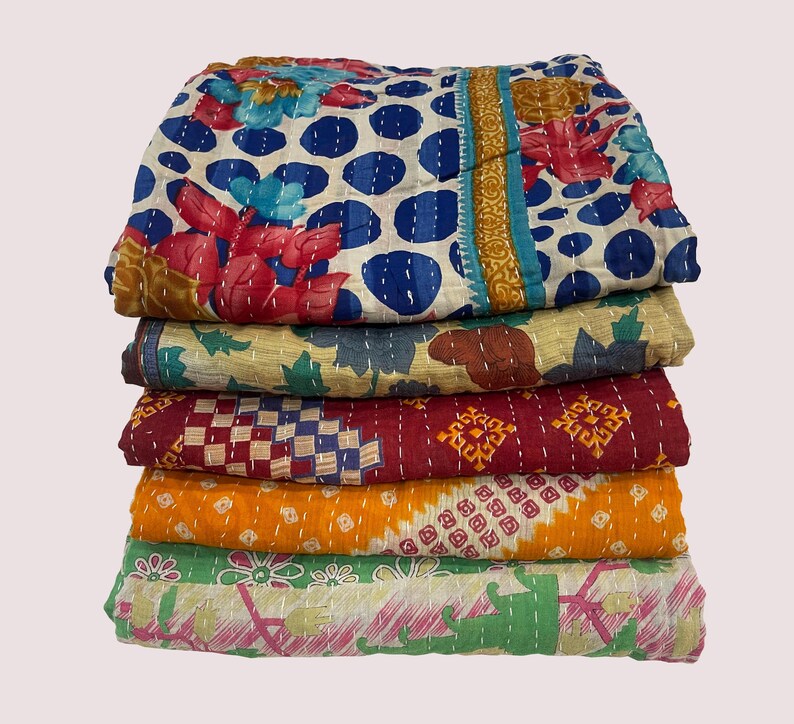 Pode incluir: Uma pilha de cinco colchas coloridas e estampadas. A colcha superior apresenta um fundo branco com bolinhas azuis e desenhos florais em vermelho e turquesa. Outras colchas exibem v&aacute;rios padr&otilde;es em tons de vermelho, laranja, verde e amarelo.