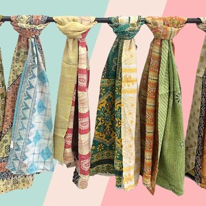 May include: A collection of ten colourful kantha scarves hanging on a rack. The scarves are made of patchwork fabric and have a variety of patterns and colours, including red, orange, yellow, green, blue, and purple.