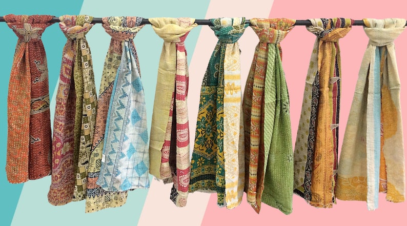 May include: A collection of ten colourful kantha scarves hanging on a rack. The scarves are made of patchwork fabric and have a variety of patterns and colours, including red, orange, yellow, green, blue, and purple.