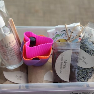 May include: A clear plastic container filled with various craft supplies, including a bottle of rubbing alcohol, a bag of dried lavender flowers, a bag of brown powder labelled "Akarnet (natural botanical dye)", and a set of pink and purple silicone moulds.