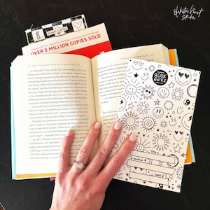 May include: An open book with text and a "Book Notes" notebook featuring sun and smiley face doodles. A bookmark with a pineapple design is visible. The book is next to a book with "OVER 5 MILLION COPIES SOLD" on the cover.