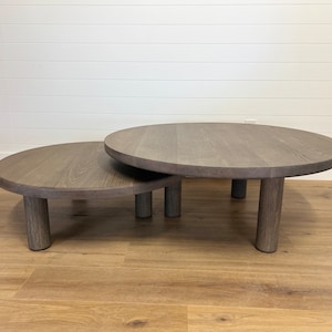 May include: A set of two round nesting coffee tables with a gray wood finish and thick, cylindrical legs.