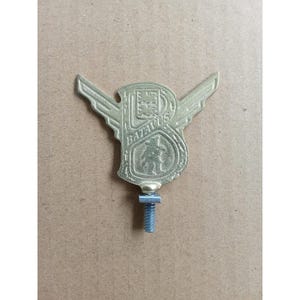 Può includere: Un emblema in metallo color oro raffigurante una lettera B stilizzata con ali che si estendono dai lati. L'emblema include il testo in rilievo BATAVUS e una piccola figura all'interno della lettera. Un bullone filettato sporge dalla base.